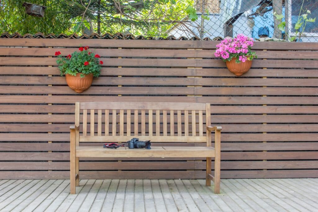 Houten tuinbank tegen een houten schutting met bloempotten, hout dat net als een tuinkist acaciahout onderhoud nodig heeft